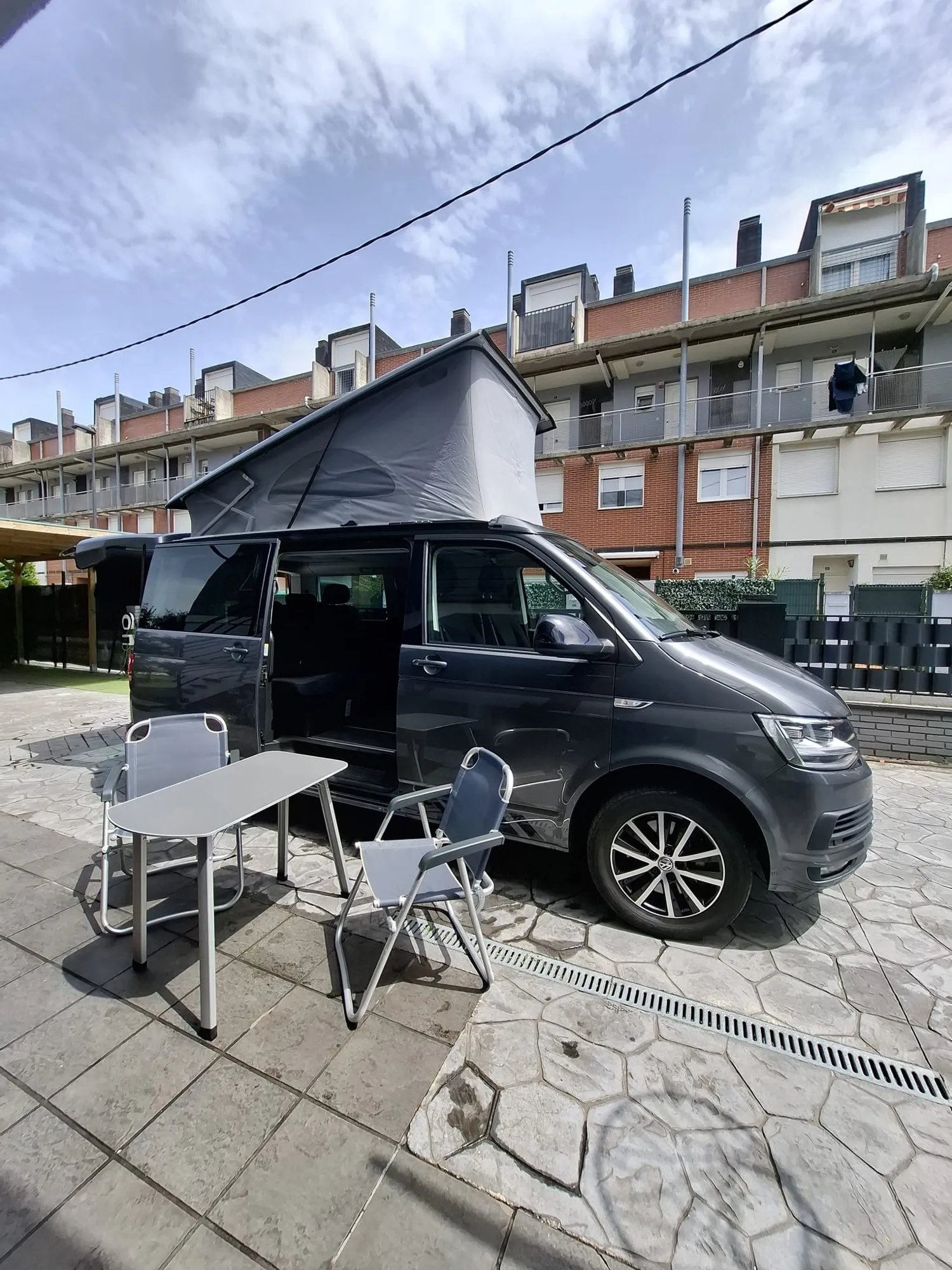 Furgoneta Volkswagen gris con el techo elevable abierto, una mesa y sillas de camping